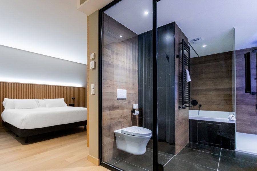 Baño moderno con ducha acristalada, paredes oscuras y cama doble visible al fondo